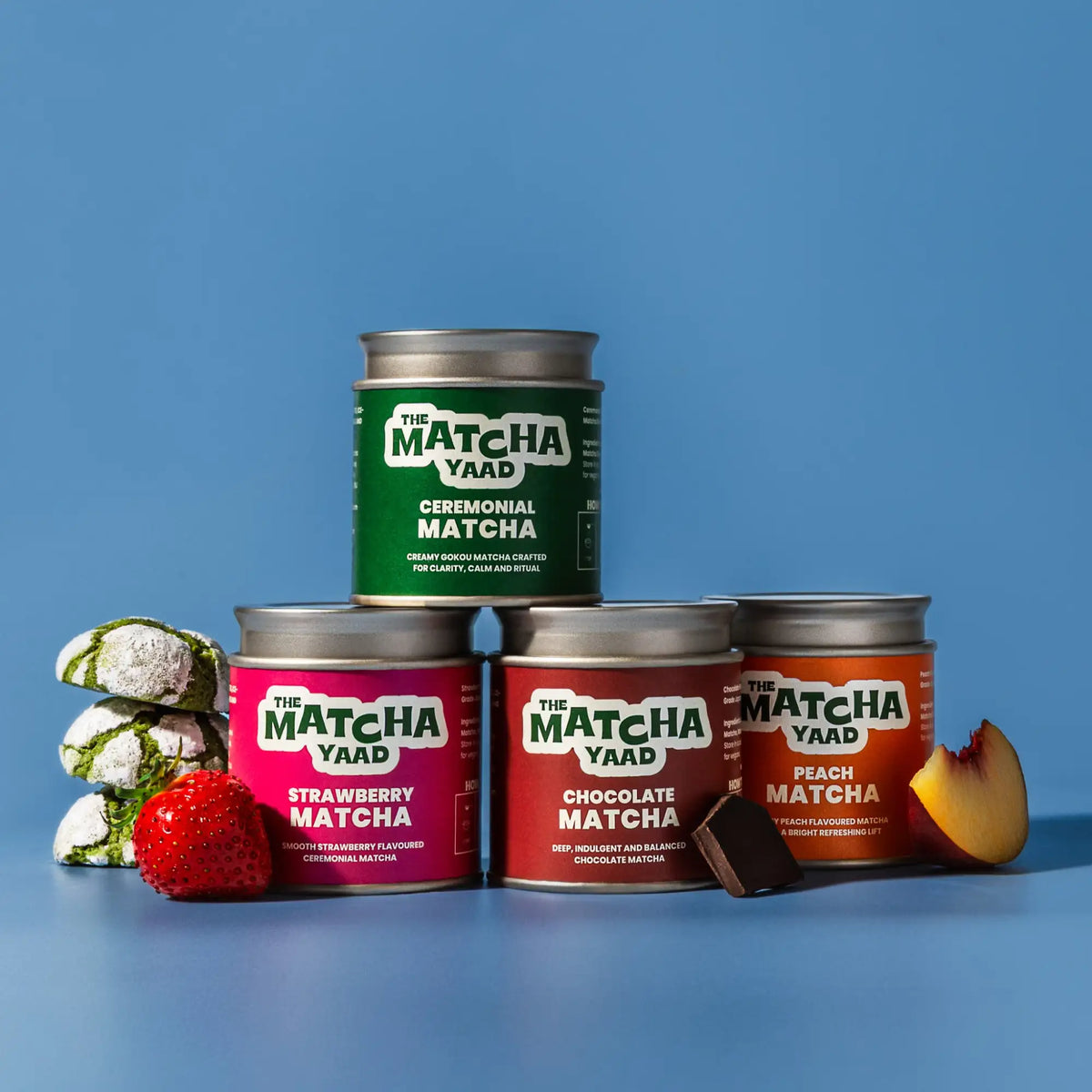 Four cylindrical metal tins of the matcha yaad matcha, each with a distinct colored label, green for ceremonial, pink for strawberry, red for chocolate, and orange for peach, and arranged in a pyramid shape.