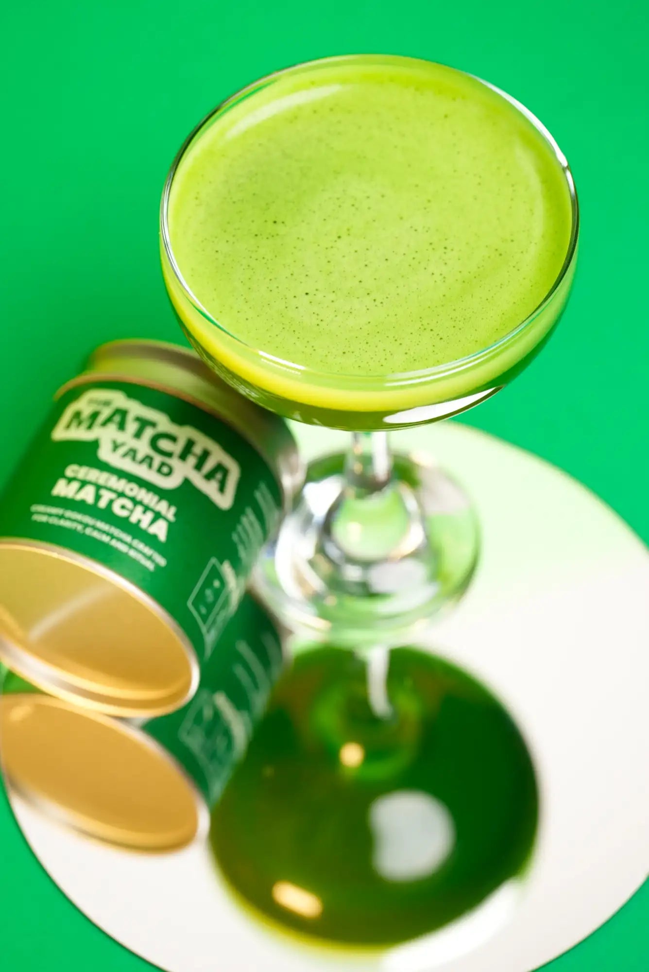 A vibrant green matcha drink served in a clear stemmed glass, accompanied by a can of ceremonial matcha powder from the brand ’matcha yaad.’